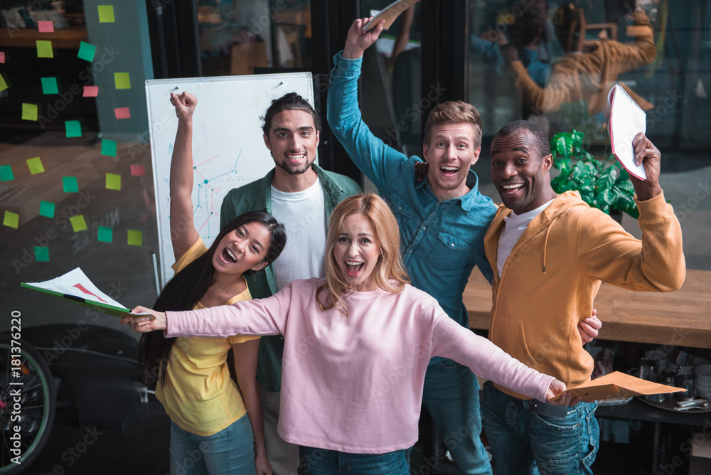 Fototapeta premium Best team ever. Top view portrait of extremely happy multinational coworkers are standing in office and screaming while expressing gladness. They are holding folders and looking at camera with joy
