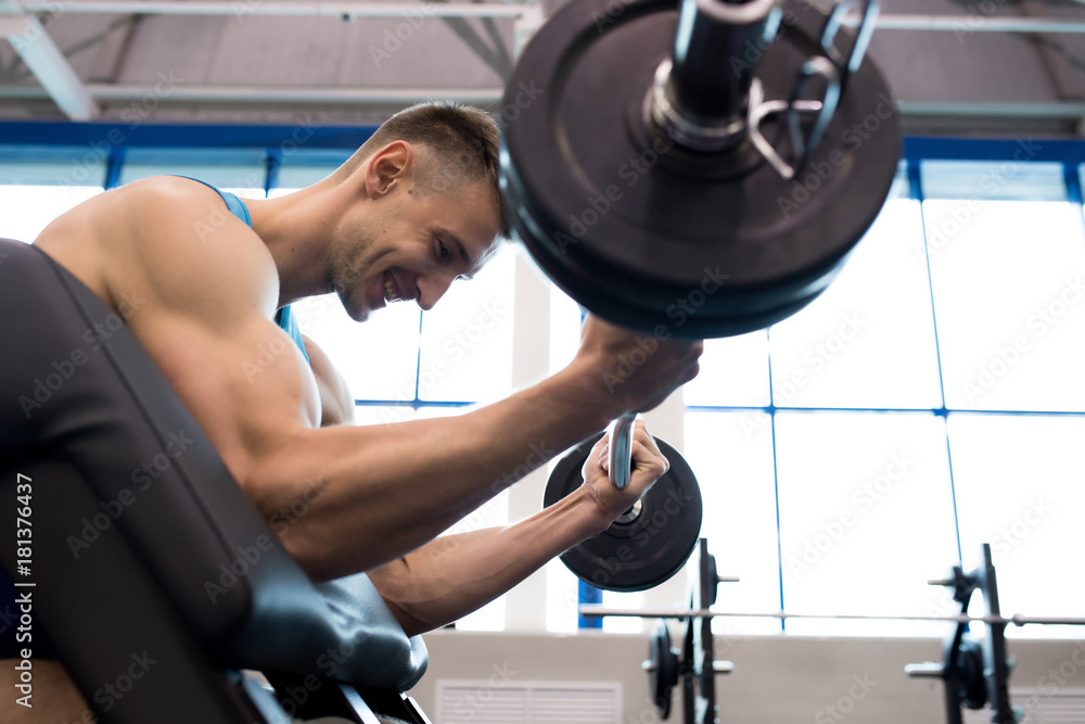 Side view portrait of handsome young man lifting barbell pumping arm muscles during strength training in modern gym, copy space Side view portrait of handsome young man lifting barbell pumping arm muscles during strength training in modern gym, copy space