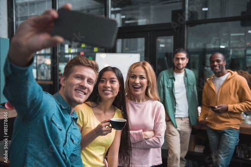 Say cheese. Joyful colleagues are posing for selfie photos while standing in office. Smiling man is using phone while asian woman is holding cup of coffee. Cute guys are leaning on table in background