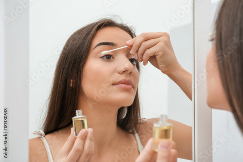 Beautiful young woman applying oil to eyelashes near mirror