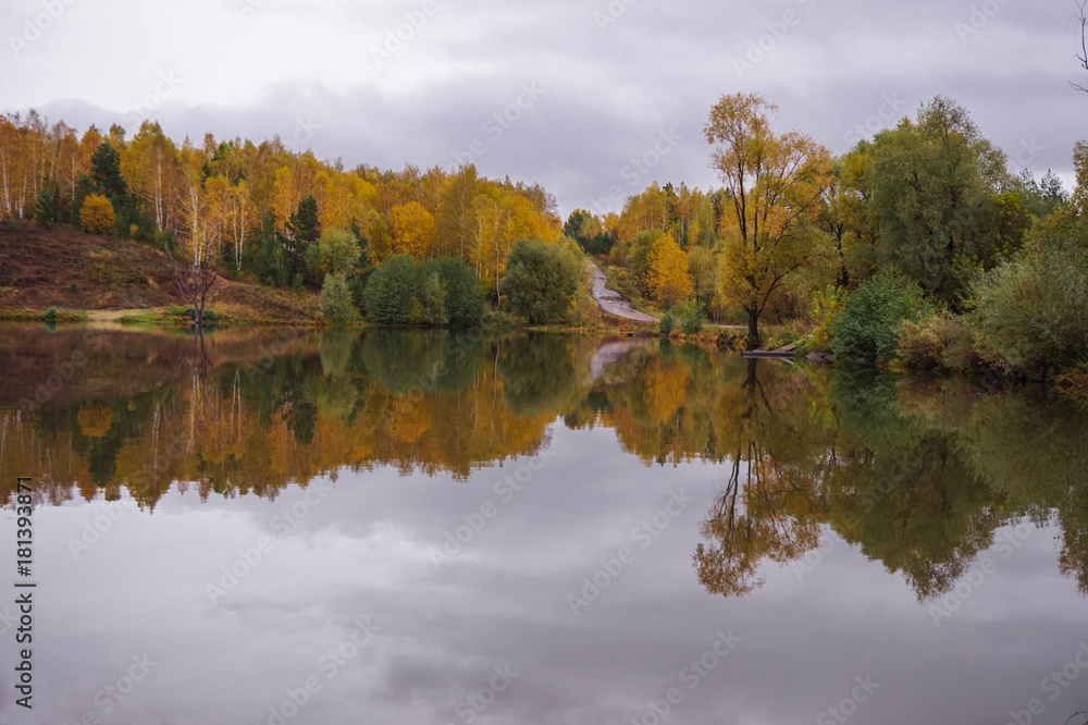The autumn forest and the road on the hills are reflected in the calm water of the forest lake.