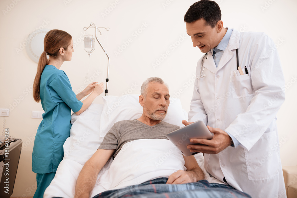The old man lies on a cot in medical ward, and next to him stands a doctor. He is showing something to old man on tablet The old man lies on a cot in medical ward, and next to him stands a doctor. He is showing something to old man on tablet