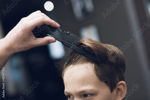 The hairdresser makes a fashionable pretty hairstyle for the boy in a modern barbershop.