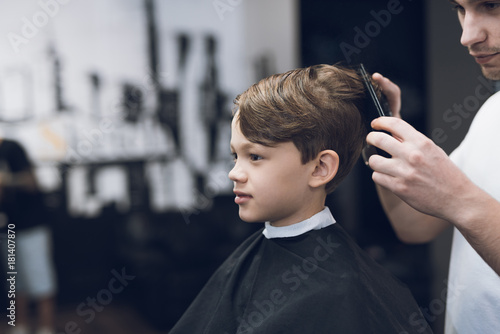 The hairdresser makes a fashionable pretty hairstyle for the boy in a modern barbershop.