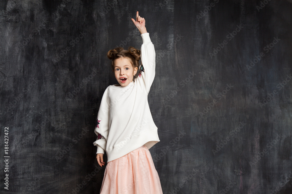 Surprised little girl in an blank white sweatshirt and pink skirt shows an index finger up against a school chalkboard.