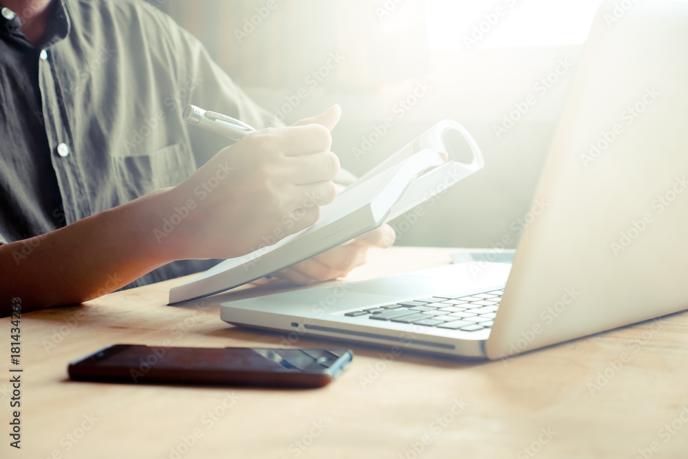 Close up of casual man writing note and using laptop on the desk, toned with sunlight Close up of casual man writing note and using laptop on the desk, toned with sunlight
