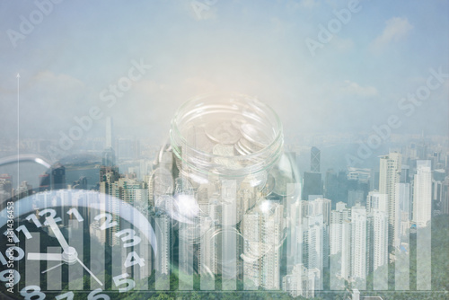 Double exposure of alarm clock and coin in glass jar with city background and graph, time for savings money concept.