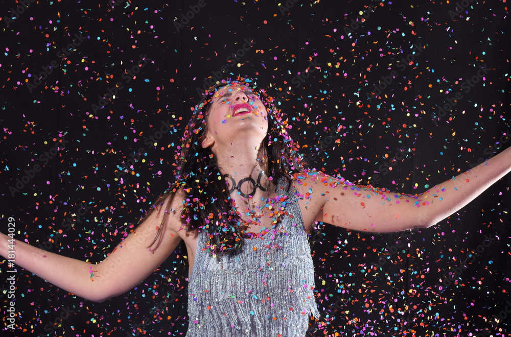 A young girl stands in front of a black background and is surrounded by confetti explosions. She laughs and smiles in her role as the party girl. A young girl stands in front of a black background and is surrounded by confetti explosions. She laughs and smiles in her role as the party girl.