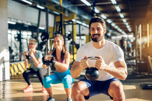 Obraz Attractive handsome bearded man holding kettlebell and doing squats in a fitness group with two girls in the modern gym.