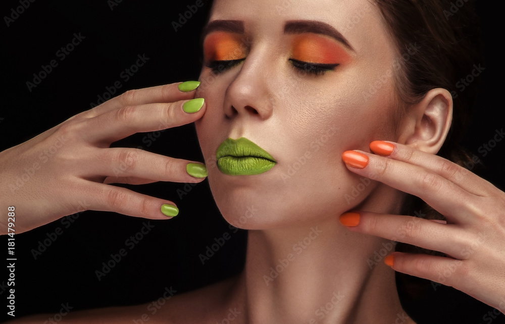 Bright colorful makeup in vivid colors on young attractive womens face. Beauty, portrait with orange eyeshadows and green lips. Sensual posing with hands. Cosmetics, healthy skin.