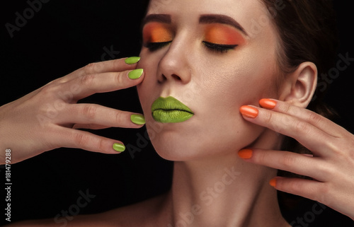 Obraz na płótnie Bright colorful makeup in vivid colors on young attractive womens face. Beauty, portrait with orange eyeshadows and green lips. Sensual posing with hands. Cosmetics, healthy skin. w salonie