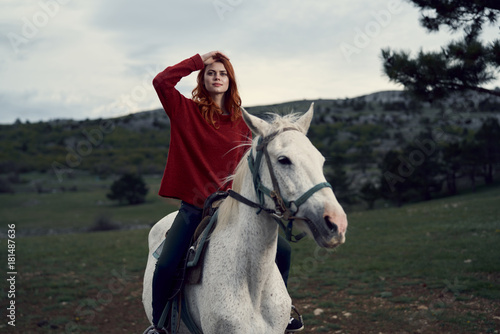 Beautiful young woman in a red sweater riding a horse on a white horse in the mountains