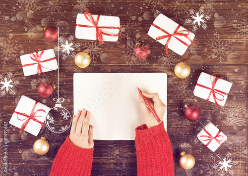 Female hands writing christmas gift list in notebook on wooden background with gifts and red and gold boiis. Toned picture with snow effect. photo