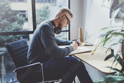 Skilled designer caucasian man drawing abstract sketch with pen.Art work process.Creative hobby.Noting ideas in copy book on workspace desk with papers in modern studio with panoramic windows.Blurred. photo