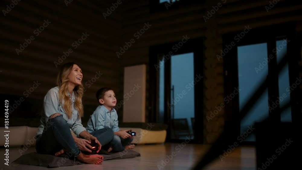 Beautiful mother with her son playing video games on gaming console in the living room of his house. Happy family in a country house. Beautiful mother with her son playing video games on gaming console in the living room of his house. Happy family in a country house.