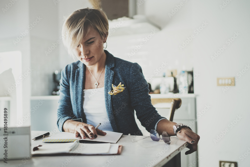 Beautiful young businesswoman writing something in note pad while sitting on armchair at living room.Charming female studying doing homework at home.Horizontal.Blurred background. Beautiful young businesswoman writing something in note pad while sitting on armchair at living room.Charming female studying doing homework at home.Horizontal.Blurred background.