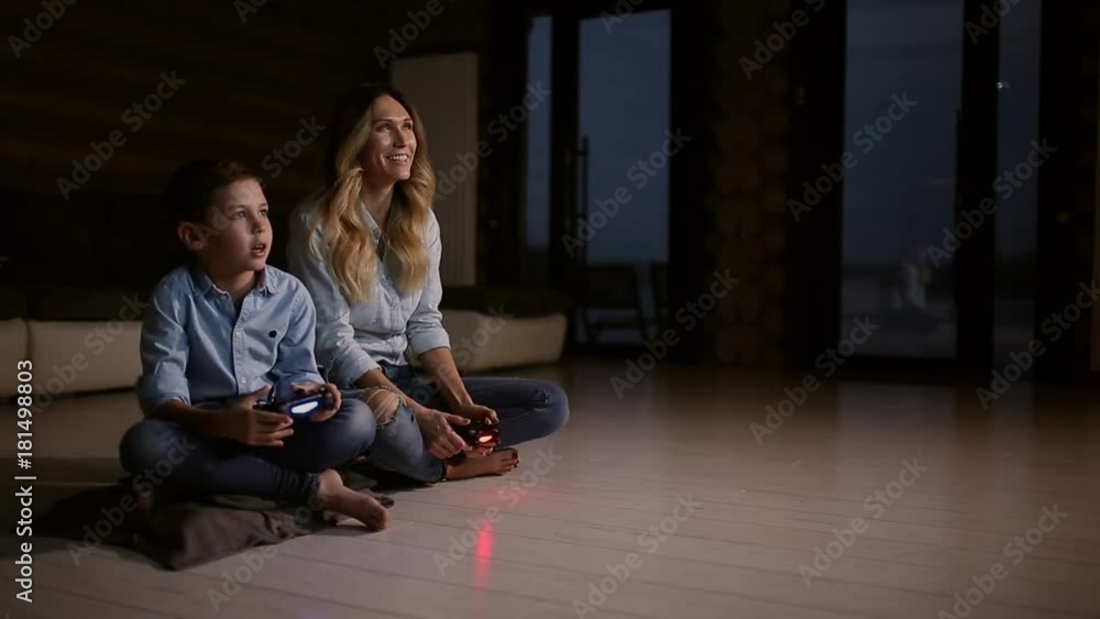 Beautiful mother with her son playing video games on gaming console in the living room of his house. Happy family in a country house. Beautiful mother with her son playing video games on gaming console in the living room of his house. Happy family in a country house.