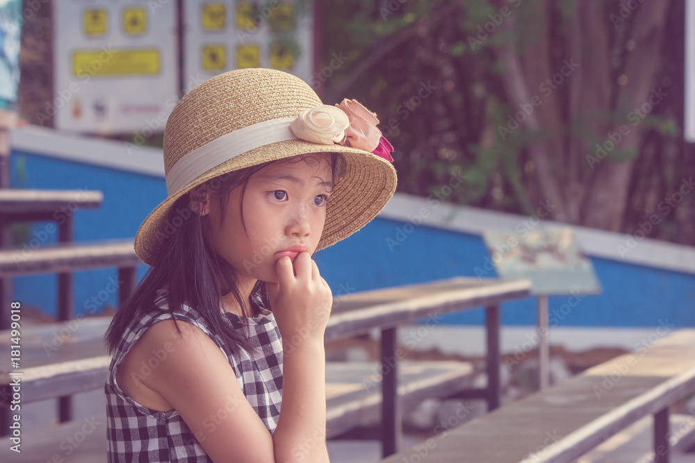 Beautiful portrait of lonely Asian cute children wearing weave hat and sitting on wooden long chair, she thinking and looking forward to something.