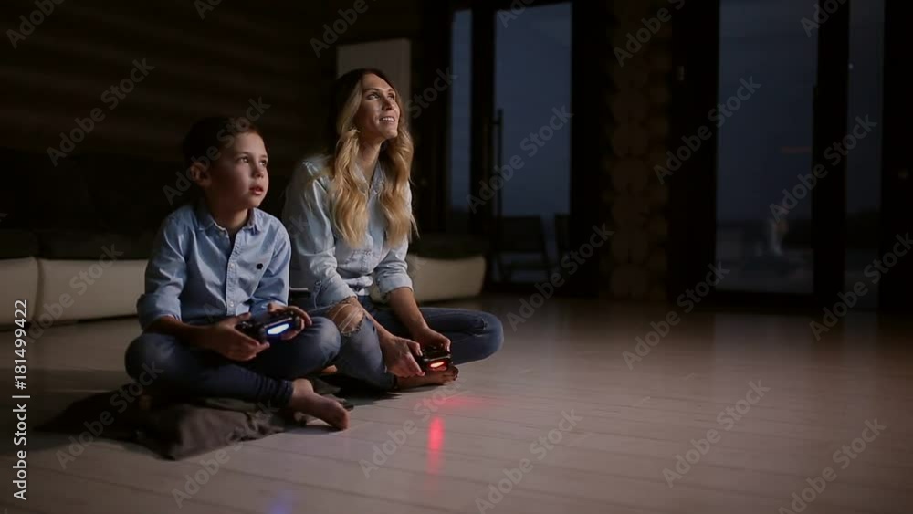 Beautiful mother with her son playing video games on gaming console in the living room of his house. Happy family in a country house. Beautiful mother with her son playing video games on gaming console in the living room of his house. Happy family in a country house.