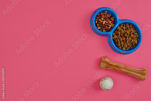 Dry pet food in bowl with a ball and dog bone on pink background top view