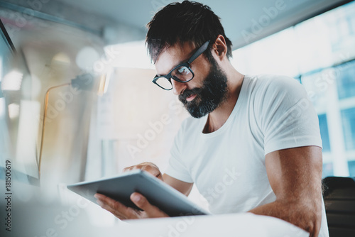 Bearded man in white tshirt wearing eye glasses and using portable electronic pro tablet computer at modern lightful office.Horizontal.Blurred background.