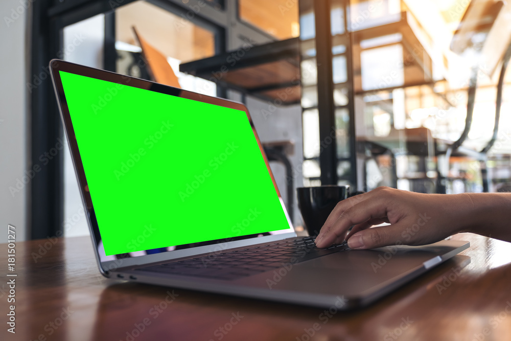 Mockup image of a hand using and typing on laptop with blank green screen and coffee cup on wooden table in cafe Mockup image of a hand using and typing on laptop with blank green screen and coffee cup on wooden table in cafe