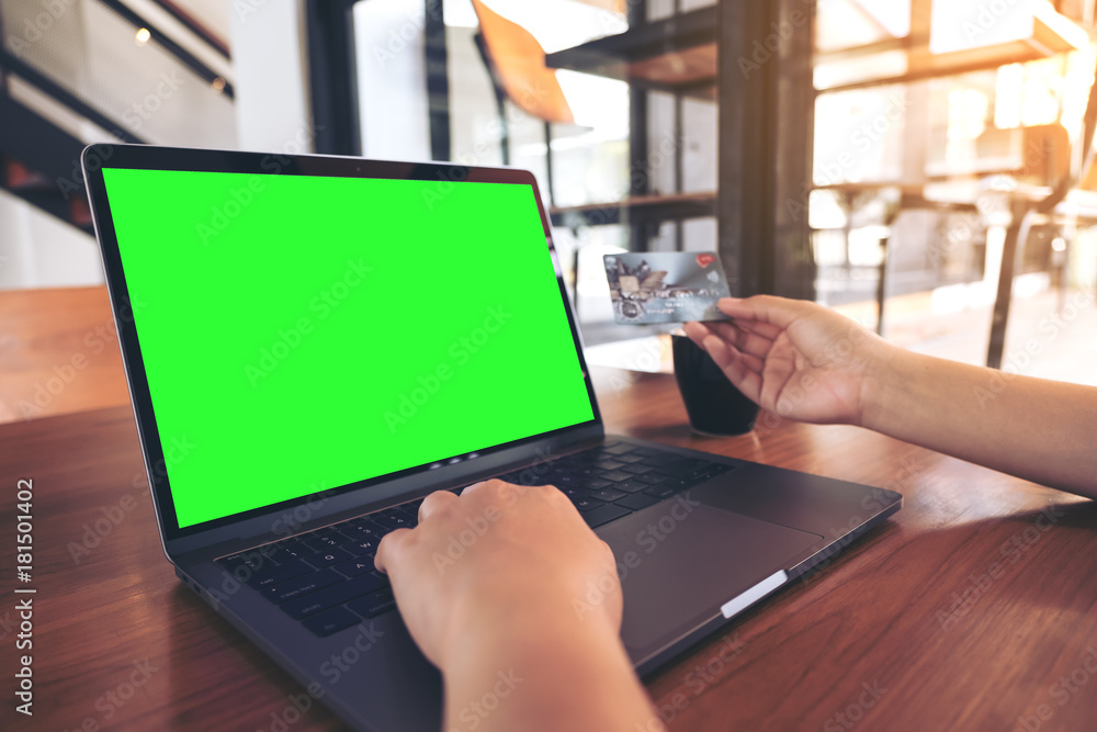 Mockup image of a hand holding credit cards while using and typing on laptop with blank green screen and coffee cup on wooden table in modern loft cafe Mockup image of a hand holding credit cards while using and typing on laptop with blank green screen and coffee cup on wooden table in modern loft cafe