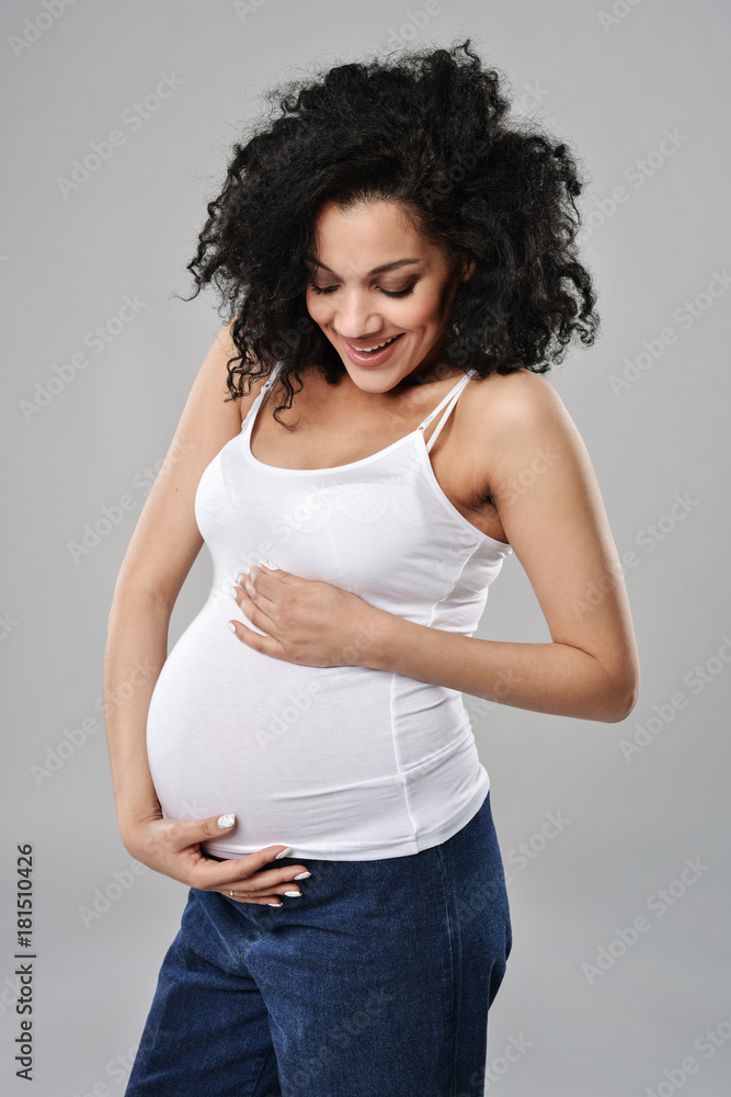 Pregnant woman smiling looking down at her tummy feeling happiness, over grey background Pregnant woman smiling looking down at her tummy feeling happiness, over grey background