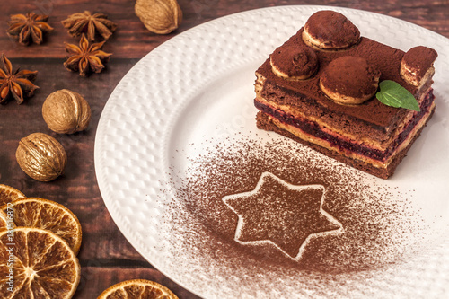 A piece of tiramisu on a white plate with a star with cinnamon, walnuts, dried oranges and anise