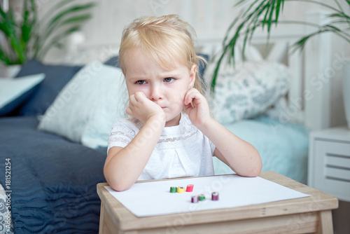 Foto Sulky grumpy attractive little blond girl sitting with paper and crayons at a small wooden desk staring off to the side with her chin on her hand