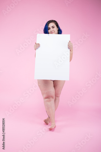 naked plus sized woman holding a white empty board in front of her in front of an even pink paper background