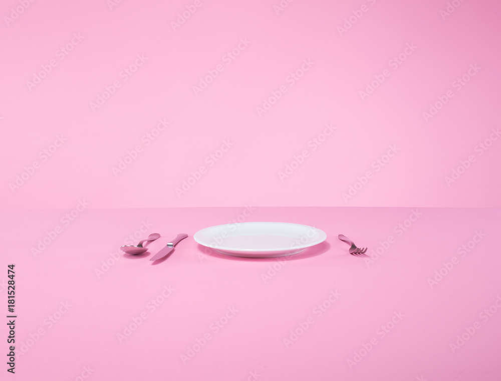 white plate, fork, spoon, and knife on a pink table in an abstract studio setting