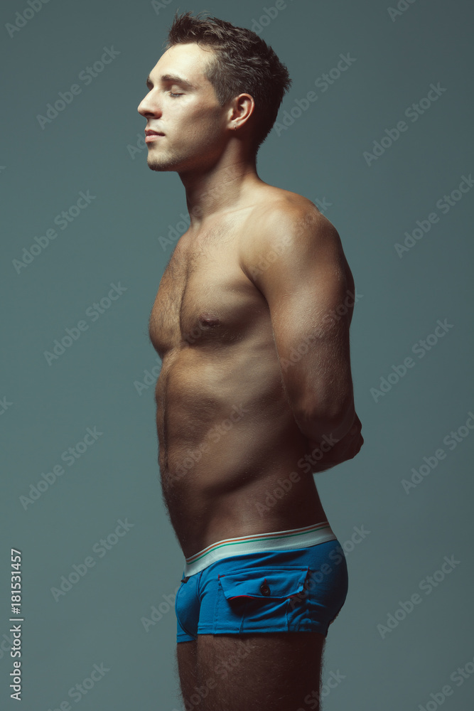 Fototapeta premium Male underwear fashion concept. Emotive portrait of young man in blue briefs posing with hands behind muscular body over blue background. Sportive style. Studio shot