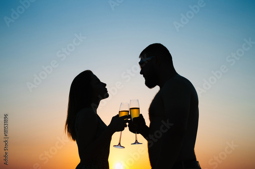 silhouettes of man and woman at sunset dramatic sky background, couple toasting wine glasses in romantic date setting, looking each other, smiling and holding in their hands glasses of champagne