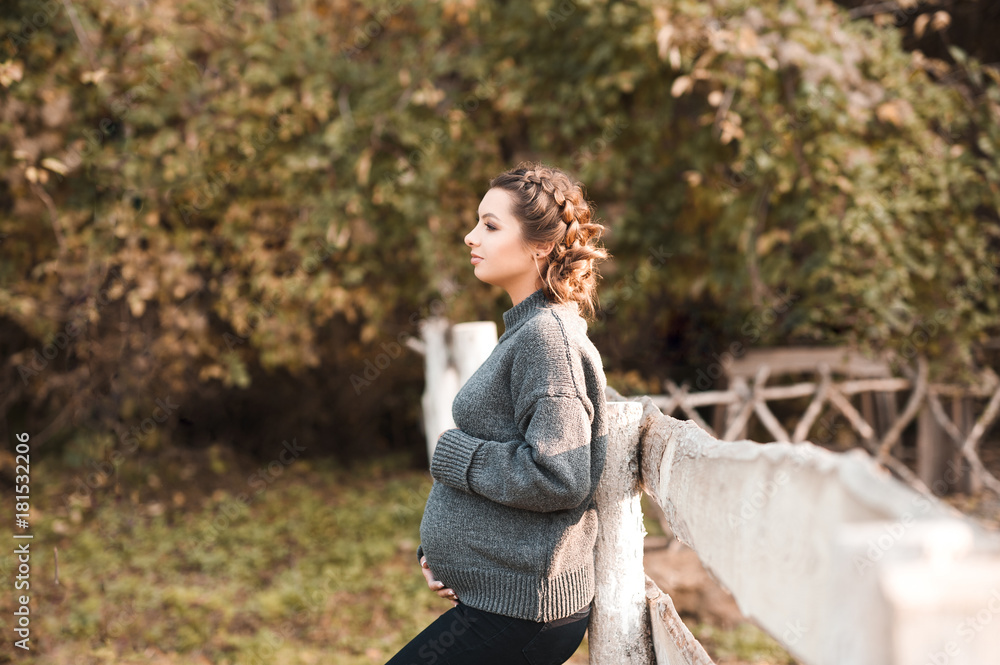 Pregnant woman wearing coy gray sweater leaning on wooden fence at countryside. Looking forward. Motherhood. Maternity. Pregnant woman wearing coy gray sweater leaning on wooden fence at countryside. Looking forward. Motherhood. Maternity.
