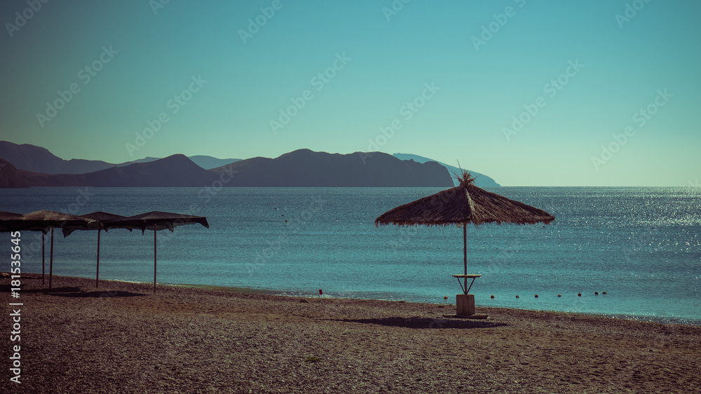 A straw beach umbrella on a pebble beach on the background of the sea and the cape. A straw beach umbrella on a pebble beach on the background of the sea and the cape.