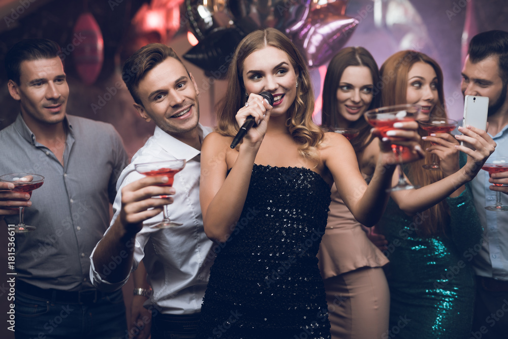 A woman in a black dress is singing songs with her friends at a karaoke club. Her friends have fun on the background.