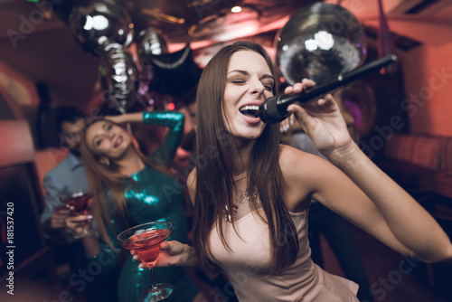 Young people have fun in a nightclub and sing in karaoke. In the foreground is a woman in a beige dress.