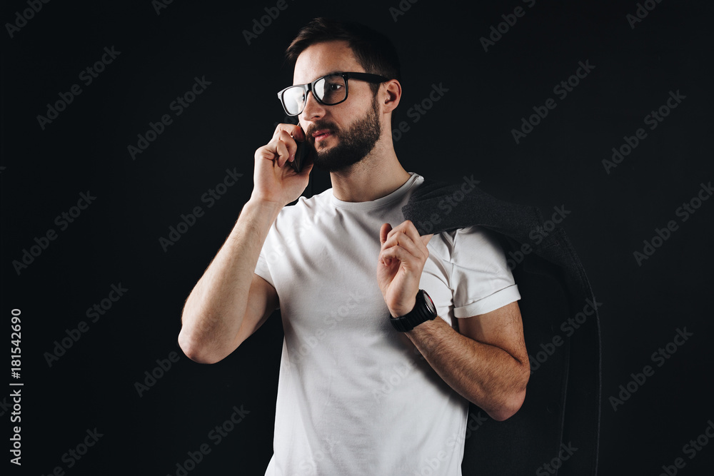 Pensive bearded young man wearing glasses and white blank tshirt talking on smartphone and holding coat in his hand. Beautiful hipster wearing in t-shirt on black background Pensive bearded young man wearing glasses and white blank tshirt talking on smartphone and holding coat in his hand. Beautiful hipster wearing in t-shirt on black background
