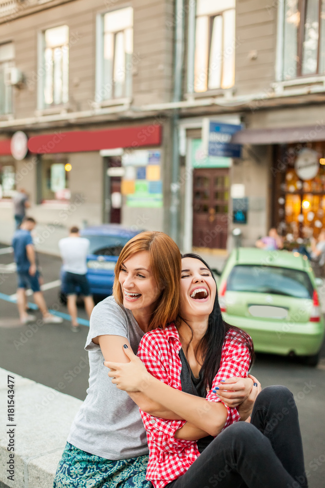 Two young girls hugging, laughting and sitting near road in the street. one brunette girl in red plaid shirt, another redhead girl wearing gray shirt and blue skirt. concept of sincere friendship Two young girls hugging, laughting and sitting near road in the street. one brunette girl in red plaid shirt, another redhead girl wearing gray shirt and blue skirt. concept of sincere friendship