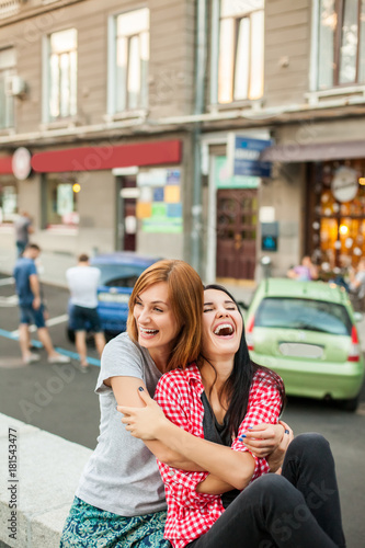 Two young girls hugging, laughting and sitting near road in the street. one brunette girl in red plaid shirt, another redhead girl wearing gray shirt and blue skirt. concept of sincere friendship
