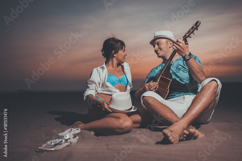 A beautiful couple enjoys a beach holiday, he playing guitar in sunset.