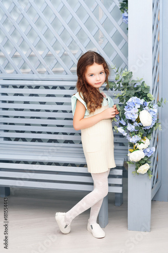 A little girl of seven years with chestnut hair, stands next to a wooden bench and a gray-blue bide sash with flowers, smiling in bright clothes, curls. The concept of a holiday.