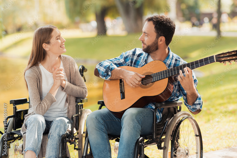 Obraz premium A man and a woman in wheelchairs met in the park. A man is playing the guitar, the woman listens and smiles.