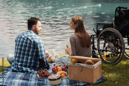 A man and a woman are sitting on the lake shore on a rug for picnics. Nearby there is a wheelchair.