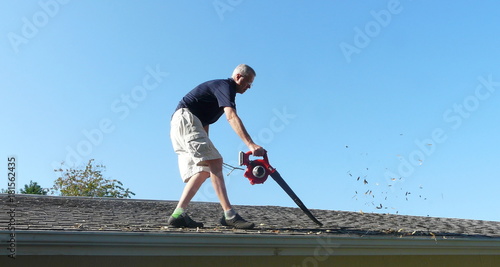 Male Homeowner Blowing Leaves from Gutter. A homeowner is on the roof of his residence using a leaf blower to remove leaves from the gutter in preparation for storm season water runoff.
