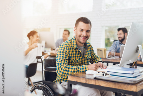 A man in a wheelchair writes with a pen in a notebook. He is working in a bright office.