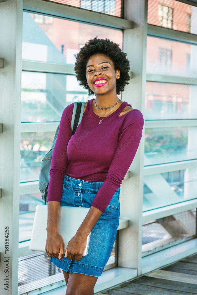 African American College Student studies in New York. Wearing long sleeve T  shirt, Denim skirt, shoulder carrying bag, holding laptop computer, girl  with short afro hairstyle,walks on campus, smiles Stock Photo |