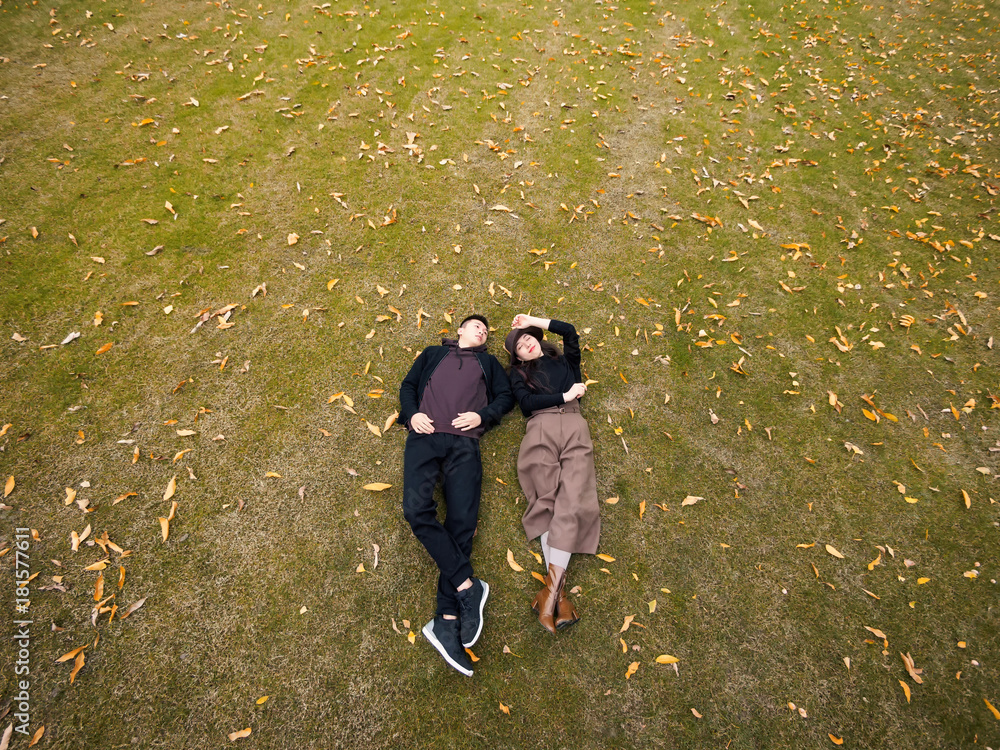 Fototapeta premium Bird's view of couple of Chinese young people laying on the grass field covered with yellow leaves in autumn day.