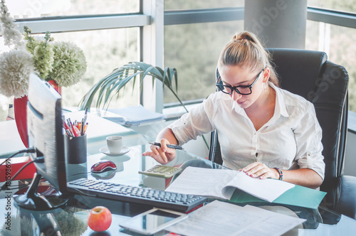 Portrait of young business lady in glasses sitting at her workplace and reads documents. Office worker at modern office workplace. Business concept. photo